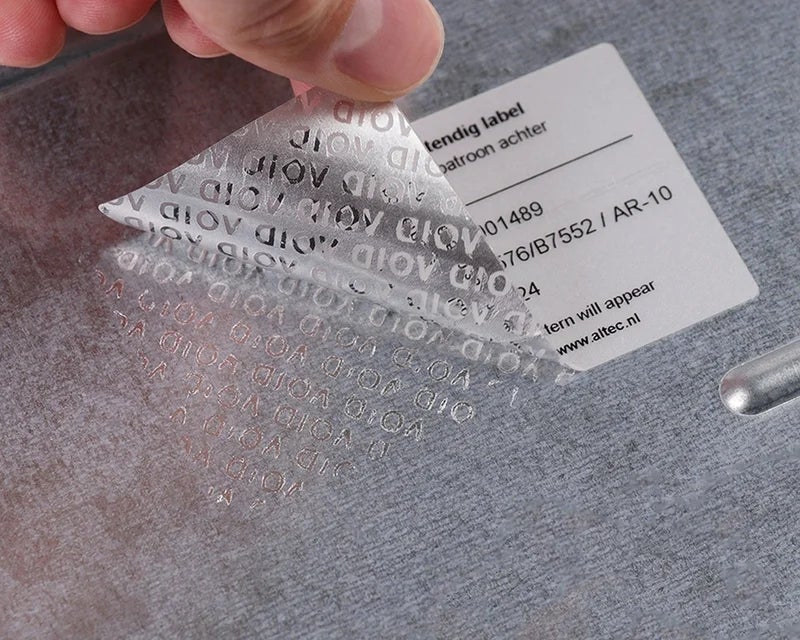 Hand verwijdert Fraudebestendig Print & Laminate label waarbij VOID-patroon zichtbaar achterblijft op oppervlak