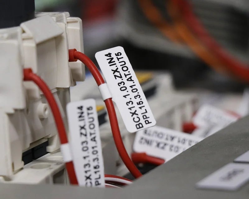 Witte vlaglabels met adercodes op rode aders bij klemmenblokken in schakelkast