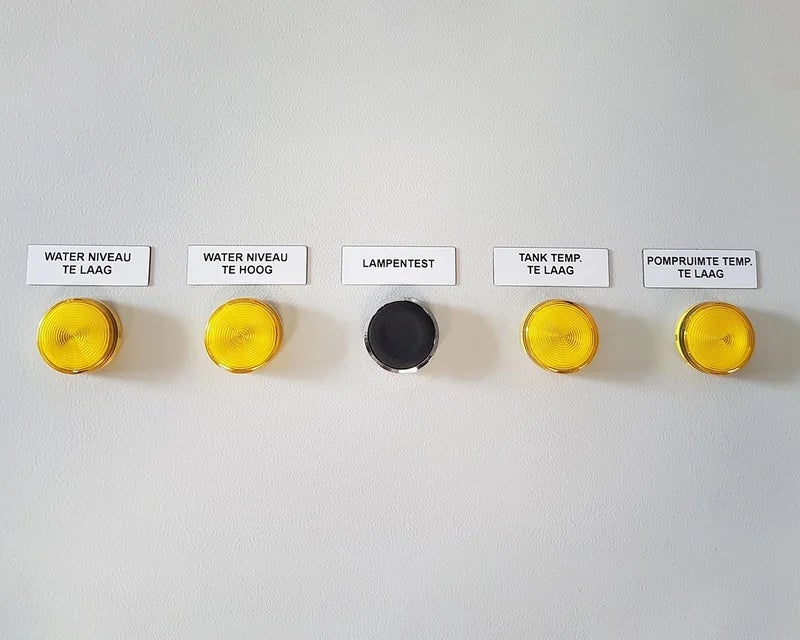Witte ResoPrint labels met omschrijvingen boven gele signaallampjes op bedieningspaneel, toegepast als alternatief voor resopal plaatjes