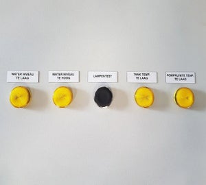 Witte ResoPrint labels met omschrijvingen boven gele signaallampjes op bedieningspaneel, toegepast als alternatief voor resopal plaatjes