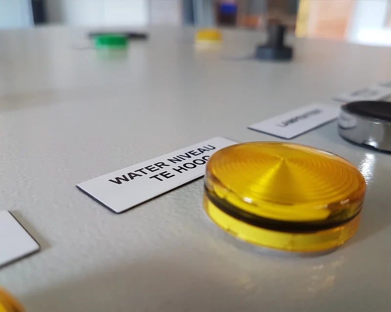 Close-up van wit ResoPrint label met omschrijving naast geel signaallampje op paneel, vergelijkbaar met een resopal plaatje