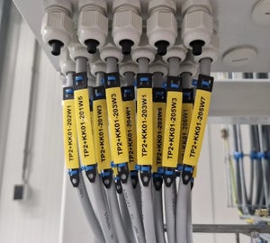 Gele krimpkousen met kabelcodes op grijze kabels bij kabelwartels in schakelkast