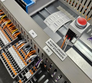 Witte ResoPrint labels met componentcodes op componenten in schakelkast naast noodstopknop, vergelijkbaar met traditionele resopals